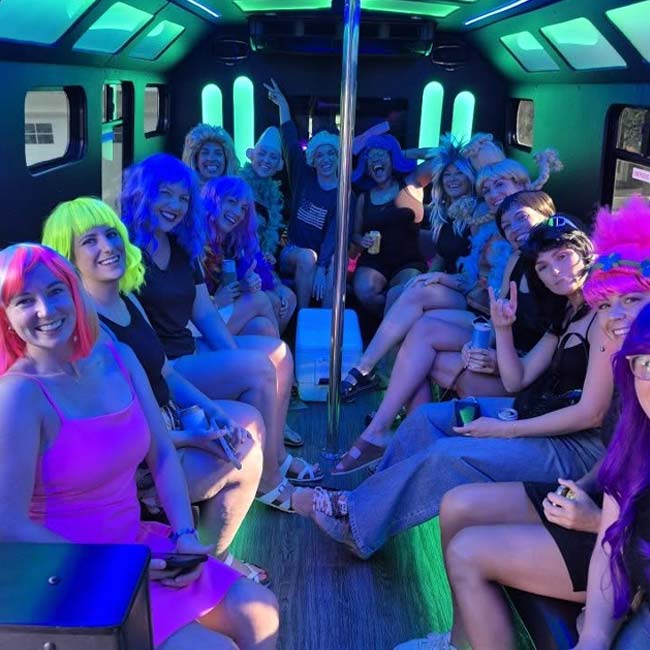Group in colorful wigs smiles in neon-lit Limo Bus, celebrating a Wisconsin bachelorette party.
