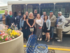A group smiles in front of a Limo Bus, with flowers and a blue chair in the Wisconsin sun.