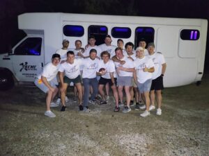 Thirteen smiling men in white shirts pose at night before an Xtreme Party Bus Service white bus.