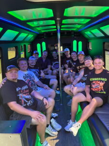 Twelve men smile inside a wedding shuttle party bus with green and blue neon lights.