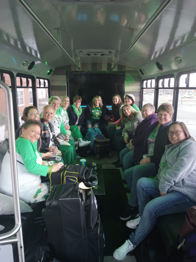 A group of women sit smiling inside a Limo Bus with bags and jackets beside them.