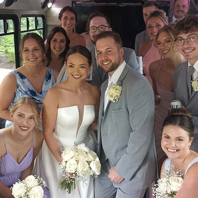 A wedding party smiles together, with a Wedding Shuttle waiting for the bride and groom outside.