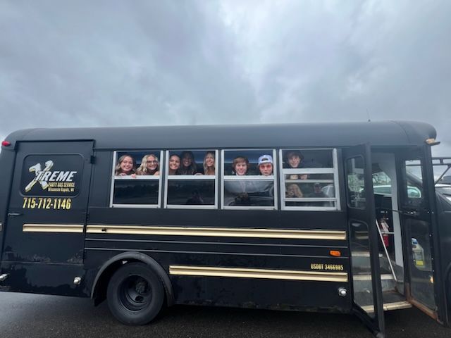 A group of smiling kids looks out from a black Wisconsin bus on a cloudy day.