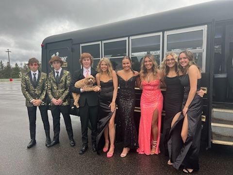 Eight teens in formal wear pose by a black party bus; one holds a small dog. It's cloudy.